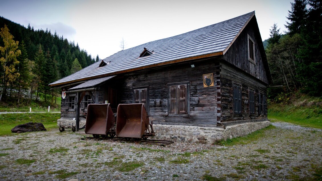 Ein traditionelles Holzhaus in einer ländlichen Umgebung, umgeben von Bäumen. Vor dem Haus stehen zwei Schaufeln, die für landwirtschaftliche Arbeiten genutzt werden. | © Holzwelt Murau