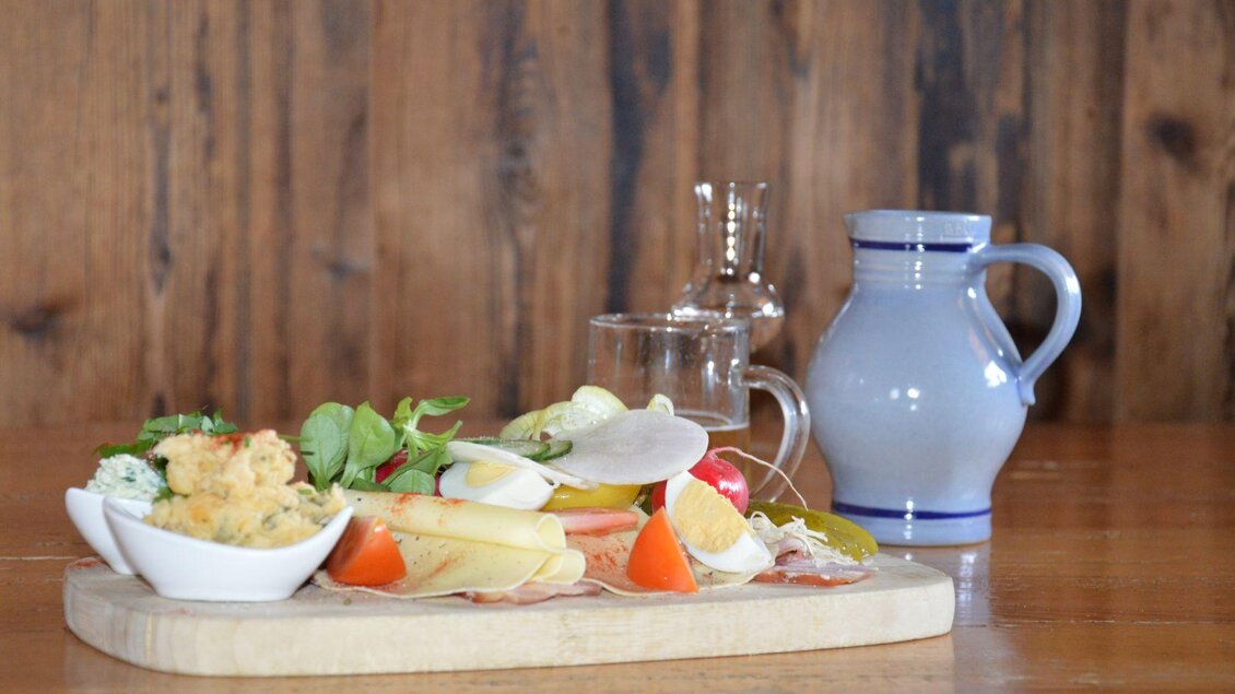 Ein Holzbrett mit einer Auswahl an frischem Gemüse, Wurst und Käse. Im Hintergrund steht eine blaue Kanne und ein Glas Wasser. | © Goasshittn