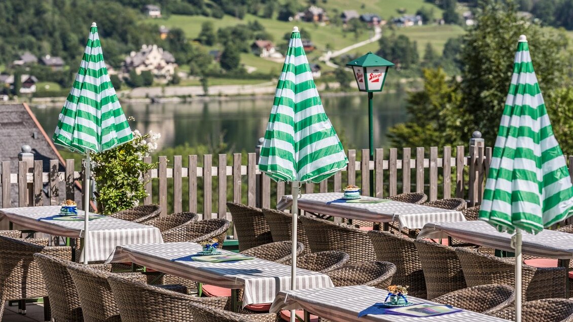 Eine gemütliche Terrasse mit Rattanmöbeln und grünen, gestreiften Sonnenschirmen. Im Hintergrund ist eine malerische Landschaft mit einem See zu sehen. | © Markus Mitterer