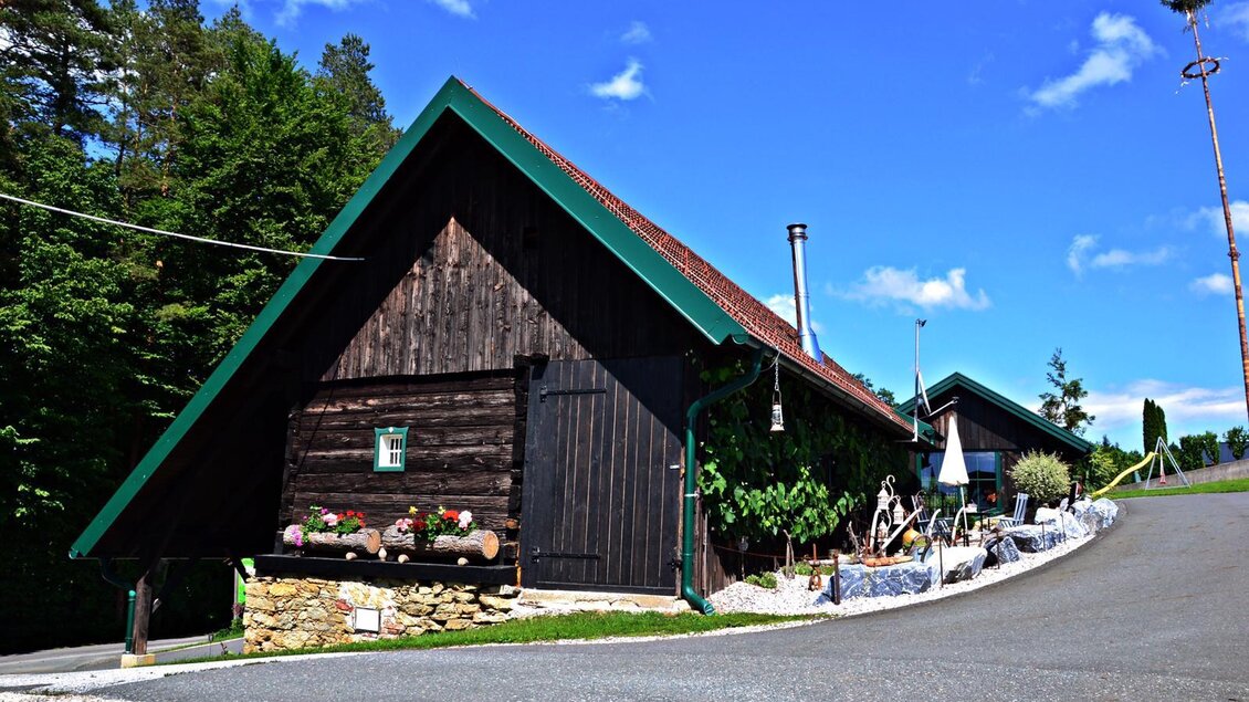 Ein charmantes Holzhaus mit einem spitzen Dach und vielen Blumen vor der Tür. Die Umgebung ist grün und die Straße ist gut gepflastert. | © Mostschank Sabines Hitt´n