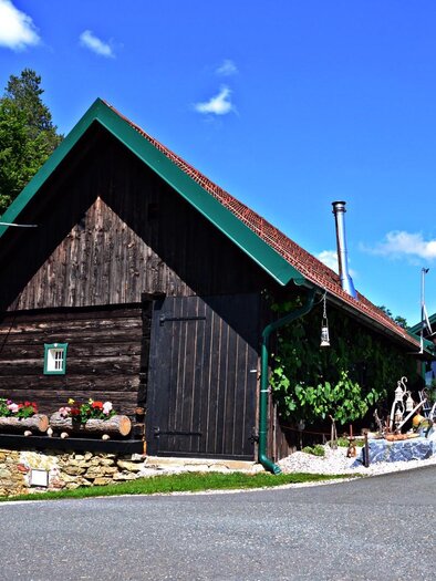 Ein charmantes Holzhaus mit einem spitzen Dach und vielen Blumen vor der Tür. Die Umgebung ist grün und die Straße ist gut gepflastert. | © Mostschank Sabines Hitt´n