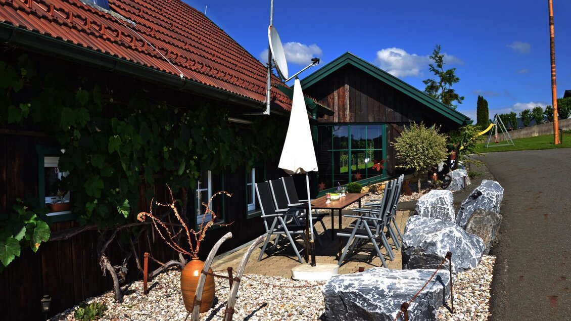 Ein gemütlicher Außenbereich mit einem Tisch und Stühlen, umgeben von großen Steinen und Pflanzen. Im Hintergrund sieht man ein Holzhaus unter blauem Himmel. | © Mostschank Sabines Hitt´n