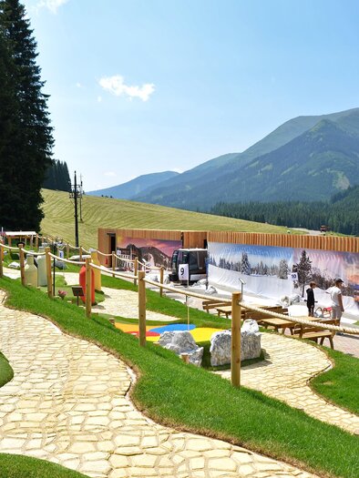 Eine malerische Landschaft mit einem Wanderweg, umgeben von Bäumen und Bergen. Im Vordergrund sind einige Spielmöglichkeiten und ein rustikales Gebäude zu sehen. | © Mountain Adventure Golf