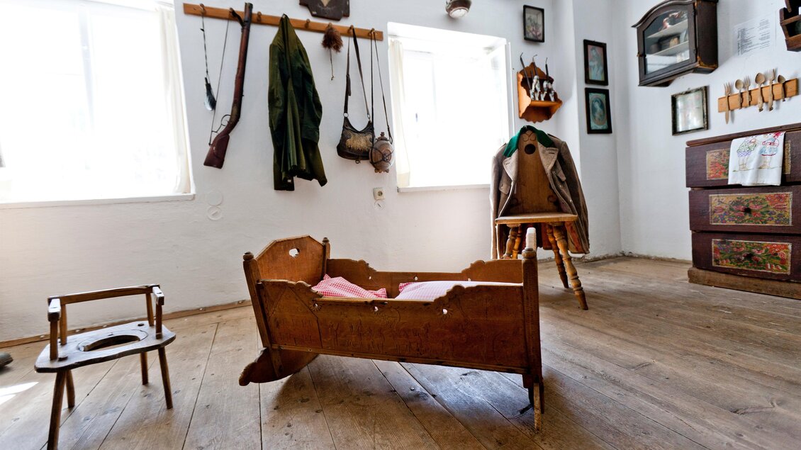 Ein gemütlicher Raum mit Holzboden und Fensterlicht. Im Vordergrund steht ein Holzkinderbett und verschiedene Gegenstände hängen an der Wand. | © Holzwelt Murau