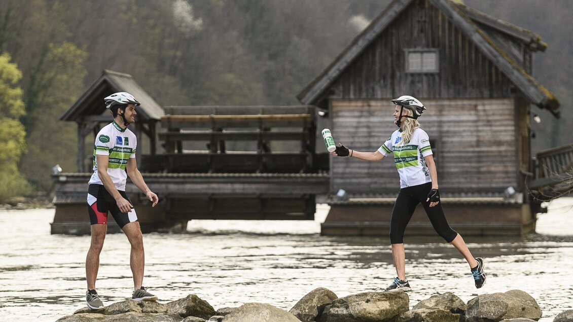 Zwei Radfahrer stehen auf Steinen im Wasser und tauschen eine Flasche aus. Im Hintergrund sind ein Holzhaus und bewaldete Hügel sichtbar. | © Thermen- & Vulkanland Steiermark_pixelmaker
