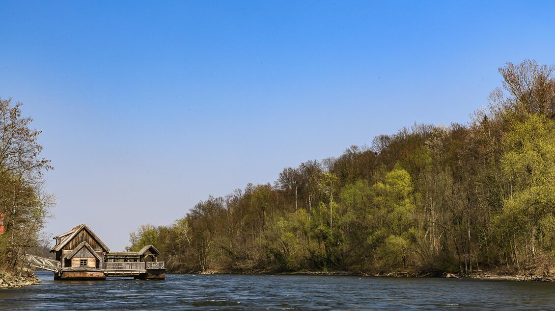 Ein ruhiger Fluss fließt durch eine grüne Landschaft. Am Ufer steht ein charmantes Holzhaus. | © Thermen- & Vulkanland Steiermark_pixelmaker