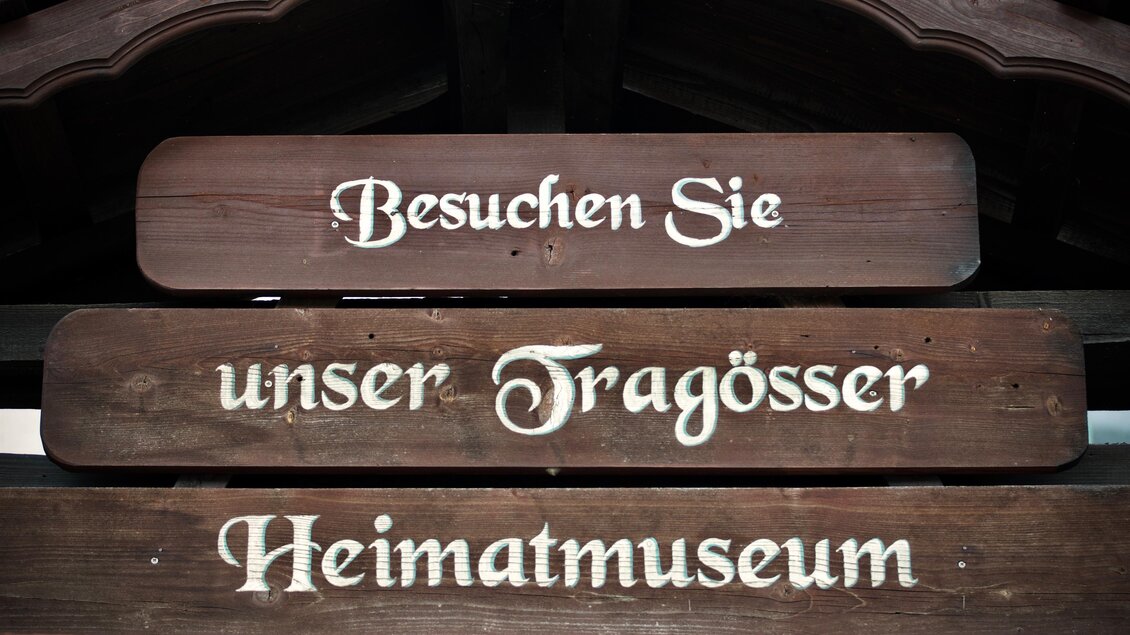 Ein Holzschild mit der Aufforderung, das Heimatmuseum in Tragöß zu besuchen. Die Beschriftung ist in geschwungener Schrift gehalten. | © TV ERZBERG LEOBEN