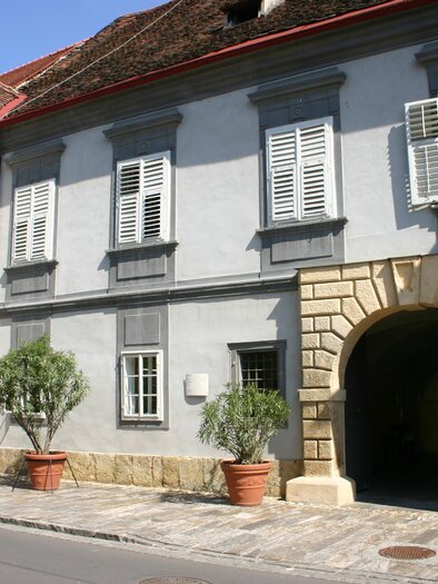 A historic building with a gray facade and large windows. It has an arch in the entrance area and is surrounded by potted plants. | © MiaZ