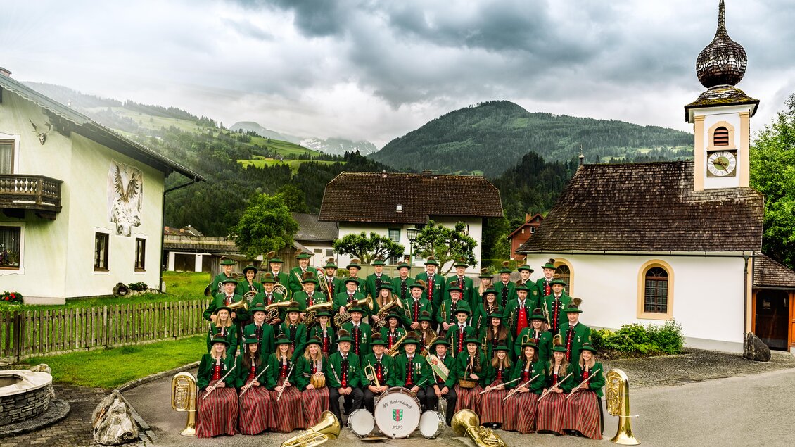 Eine große Gruppe von Musikern steht in traditioneller Tracht vor einer Kirche und malerischen Bergen. Sie halten Musikinstrumente und lächeln für das Foto.