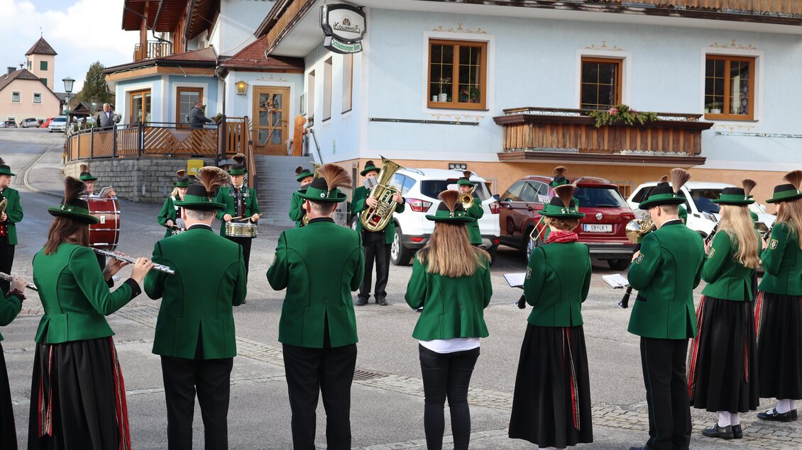 Eine Gruppe von Musikern in grünen Uniformen spielt auf einer Straße vor einem Gebäude. Im Hintergrund sind Autos und eine bayerische Landschaft zu sehen.