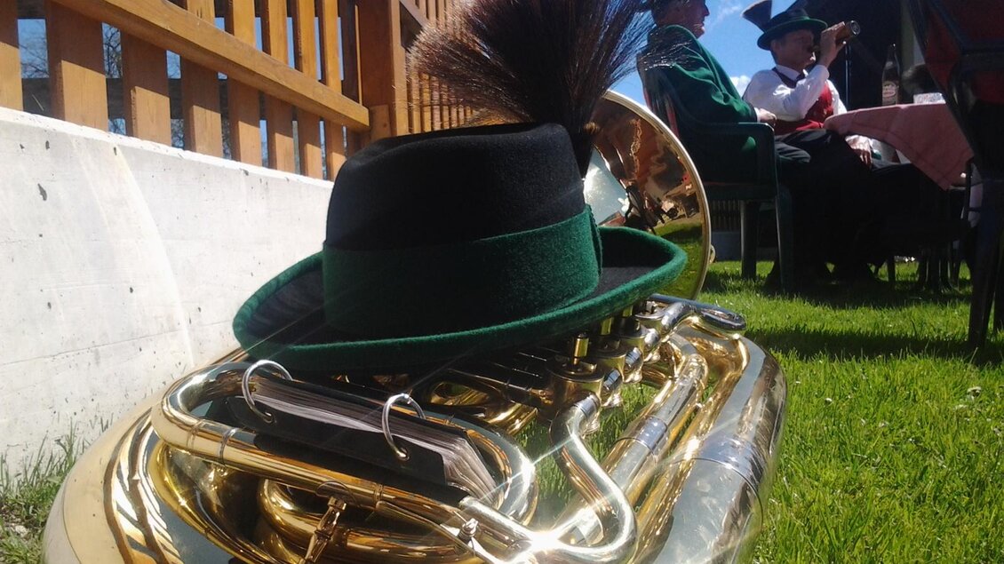 Eine glänzende Tuba liegt auf dem Gras, bedeckt mit einem traditionellen Hut. Im Hintergrund sind Personen in Trachten zu sehen.