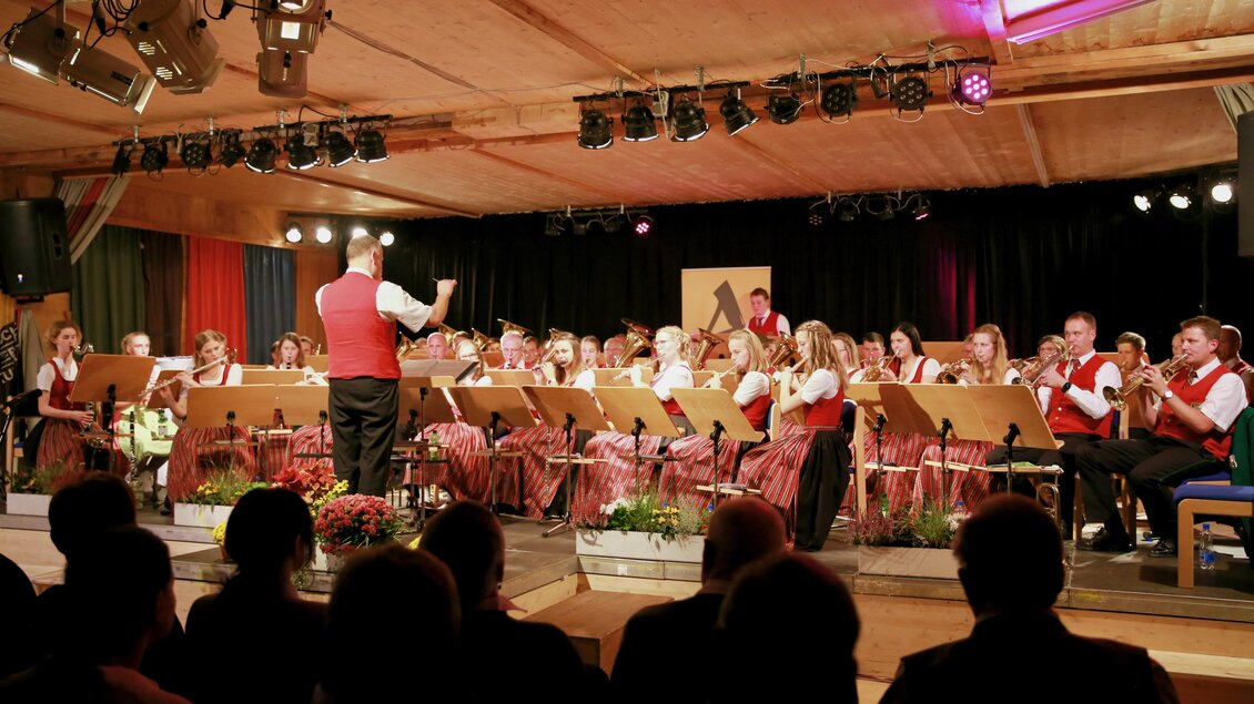 Ein Orchester spielt auf einer Bühne, geleitet von einem Dirigenten. Die Musiker tragen traditionelle Kleidung und die Atmosphäre ist festlich.