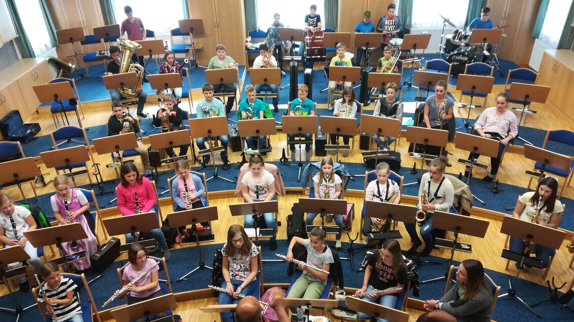 Eine Gruppe von Kindern sitzt in einem Probenraum mit Musikinstrumenten und Notenständern. Sie sind bereit, gemeinsam Musik zu machen.