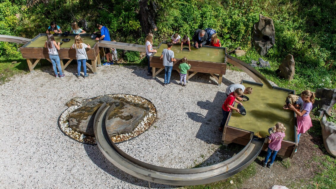 Ein Spielplatz im Freien mit mehreren Holztischen, an denen Kinder und Erwachsene aktiv sind. Im Hintergrund sind grüne Bäume und ein kleiner Wasserbereich zu sehen. | © Edelstein Kranzelbinder