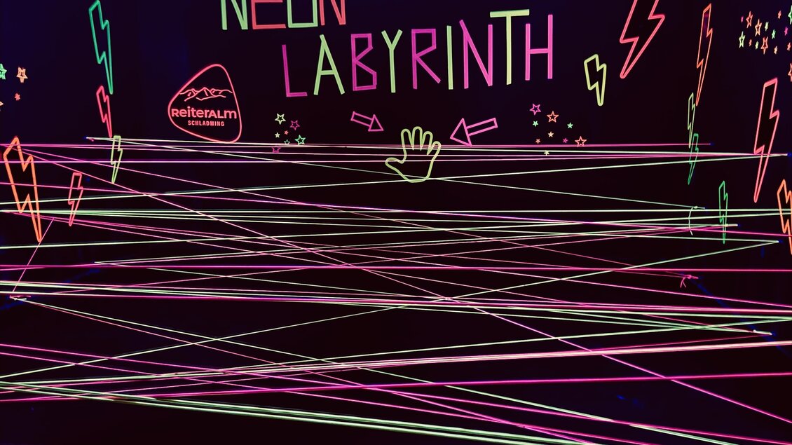 Ein neonbeleuchtetes Labyrinth mit bunten Lichtstrahlen zieht sich durch den Raum. An der Wand steht der Schriftzug „NEON LABYRINTH“. | © Reiteralm