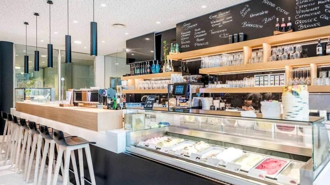 Thekenbereich von NOVA - Das Café in der Therme NOVA Köflach mit Vitrine, Barhockern und Menütafel im Hintergrund. | © Hotel & Therme NOVA