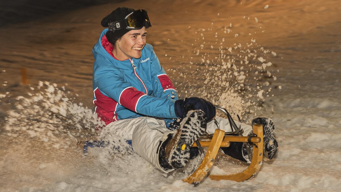 <p>Ein Junge fährt mit einem Schlitten den verschneiten Hang bei der Nachtrodelbahn Grafenwiese in Tauplitz hinunter. Die Piste ist im Hintergrund leicht beleuchtet und Schnee spritzt auf.</p> | © Melanie Fritz