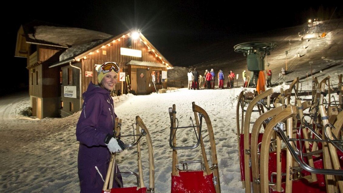 <p>Eine Frau in lila Skibekleidung steht mit einem Schlitten in der Hand vor der Skihütte Grafenwiese bei Nacht. Im Hintergrund sind beleuchtete Skifahrer und weitere Schlitten zu sehen.</p> | © Mirja Geh
