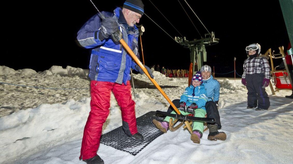 <p><p>Ein Mitarbeiter hilft zwei Rodelfahrern auf einem Schlepplift mit der Rodel. Es ist Nacht und die Umgebung ist mit Schnee bedeckt.</p></p> | © Mirja Geh