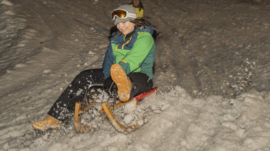 <p>Ein Mädchen fährt mit einem Schlitten die schneebedeckte Piste bei der Grafenwiese in Tauplitz hinab und hat Spaß. Im Hintergrund ist eine weitere Person zu sehen.</p>