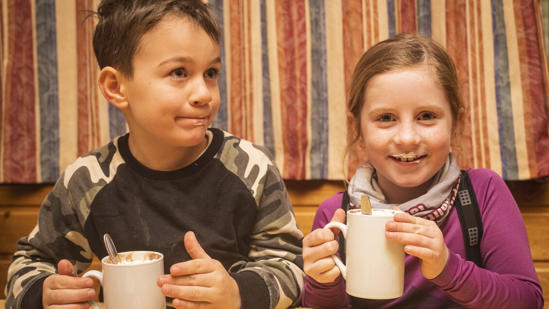 <p>Zwei Kinder sitzen nach dem Nachtrodeln bei der Grafenwiese in Tauplitz bei einer Tasse Kakao in einer warmen Hütte. Sie lächeln freundlich und scheinen die gemeinsame Zeit zu genießen.</p>