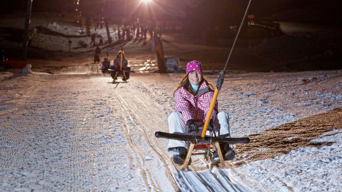 <p>Eine Frau sitzt auf einem Schlitten und fährt bei Nacht mit dem List, auf einer beleuchteten Piste den Hügel hoch. Im Hintergrund sind andere Rodler und Schneelandschaft sichtbar.</p> | © Mirja Geh