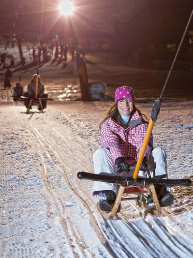 <p>A woman sits on a sled and rides at night with the list, up a lit slope. In the background, other sledders and snowy landscape are visible.</p> | © Mirja Geh
