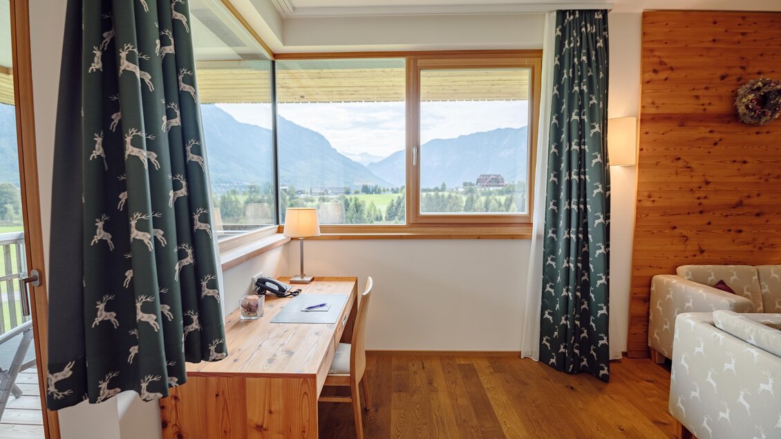 <p>Ein gemütliches Zimmer im Narzissen Vital Resort in Bad Aussee mit einem Holzschreibtisch und großen Fenstern, welche einen Blick auf die umliegenden Berge bieten. Die Vorhänge haben ein Hirsch-Muster und die Einrichtung ist modern und einladend.</p> | © Karl Steinegger