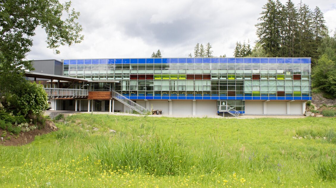 Ein modernes Gebäude mit großen Fenstern und einer bunten Fassade. Umgeben von einer grünen Wiese und Bäumen. | © Natechnikum