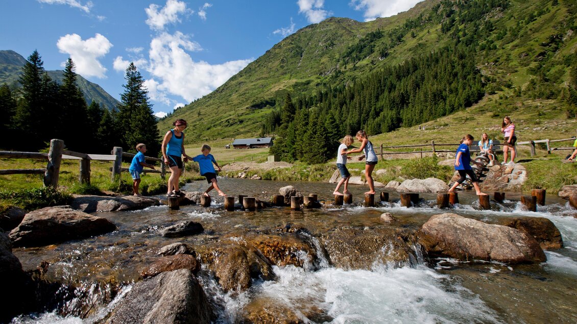 Eine Gruppe von Menschen überquert einen kleinen Bach in einer wunderschönen Berglandschaft. Die Sonne scheint, und die umliegenden Berge sind grün und schön. | © Holzwelt Murau