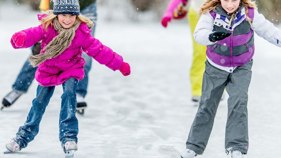 Zwei Mädchen laufen fröhlich auf einer Eisbahn. Sie tragen bunte Winterkleidung und genießen den Schnee. | © Narzissendorf Zloam www.zloam.at