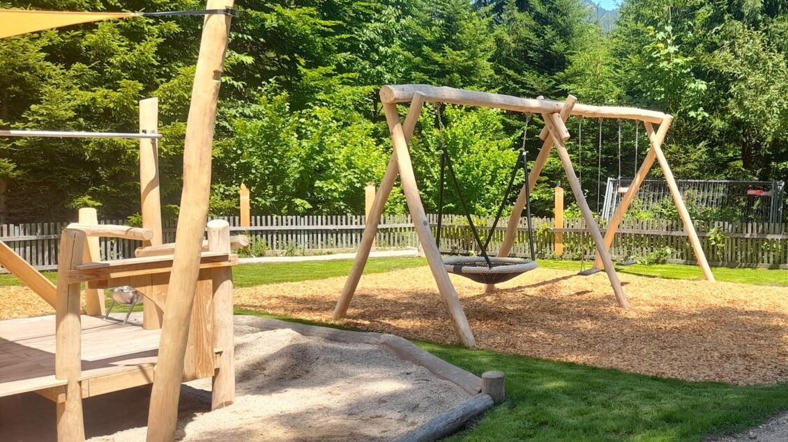 <p>Der Naturspielplatz am Ödensee mit einer Holzschaukel. Der Bereich ist von Bäumen umgeben und es gibt eine grüne Wiese.</p> | © Isabell Schachner