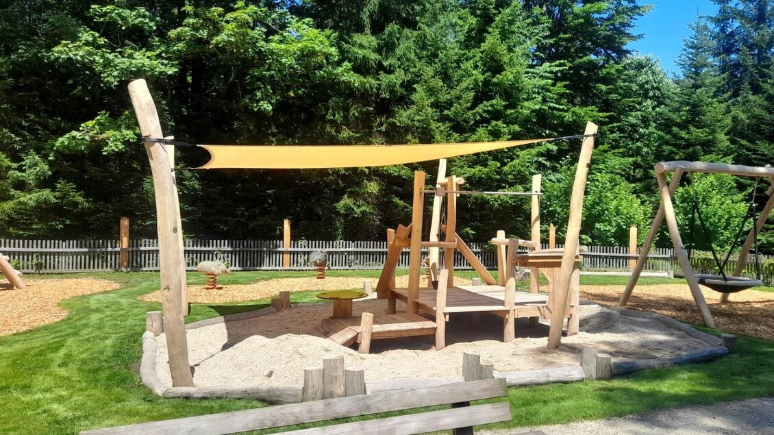 <p>Der moderne Spielplatz aus Holz mit einer Sandfläche und einem Sonnensegel am Ödensee. Im Hintergrund sieht man hohe Bäume und eine Schaukel.</p> | © Isabell Schachner