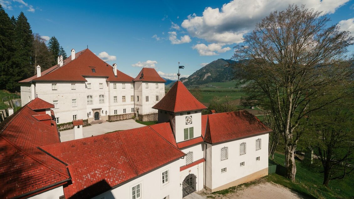 Die historische Anlage umfasst mehrere weiße Gebäude mit markanten roten Dächern. Im Hintergrund erstreckt sich eine alpine Landschaft mit Wäldern und Bergen. | © Christoph Lukas