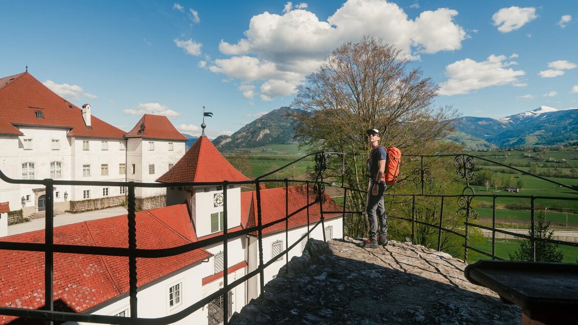 Ein Wanderer blickt von einer historischen Mauer auf Schloss Trautenfels udn das umliegende Ennstal. Im Hintergrund sind bewaldete Berge und schneebedeckte Gipfel zu sehen. | © Christoph Lukas
