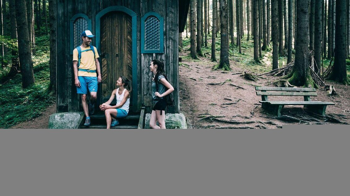 Eine Gruppe von Wanderern macht Pause vor der Kapelle Heiligenbrunn am Rundwanderweg. Das Gebäude steht in einem Nadelwald neben einer Holzbank. | © Armin Walcher