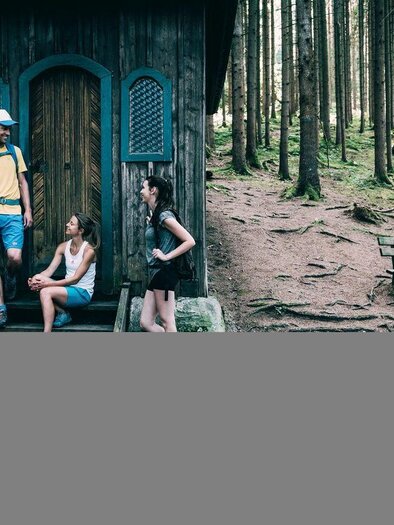 A group of hikers is taking a break in front of the Heiligenbrunn chapel on the circular hiking trail. The building is located in a coniferous forest next to a wooden bench. | © Armin Walcher