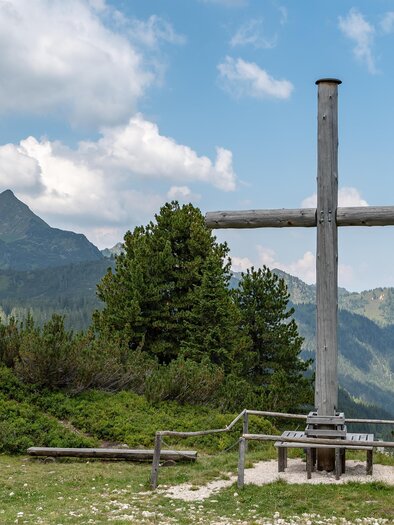 Ein großes Holzkreuz steht auf einer Bergwiese mit grünen Bäumen und einem hügeligen Panorama im Hintergrund. Der Himmel ist teils bewölkt und es gibt eine schöne Aussicht auf die Berge. | © Isiwal