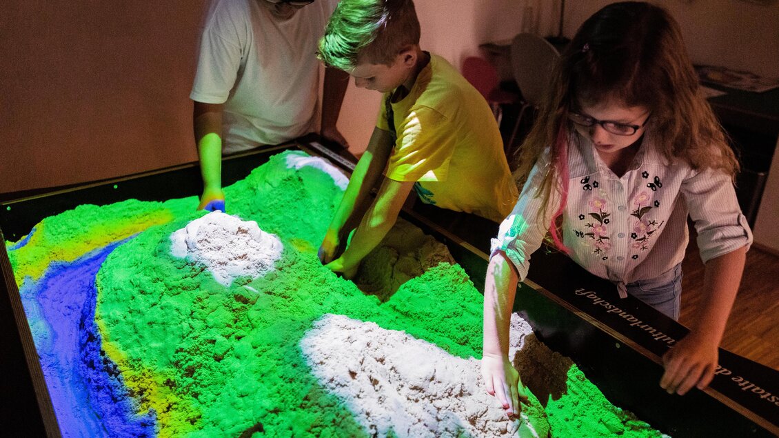 Drei Kinder spielen an einem interaktiven Sandsimulationstisch. Sie gestalten mit ihren Händen eine farbenfrohe Landschaft aus Sand und Wasser. | © TV Murau