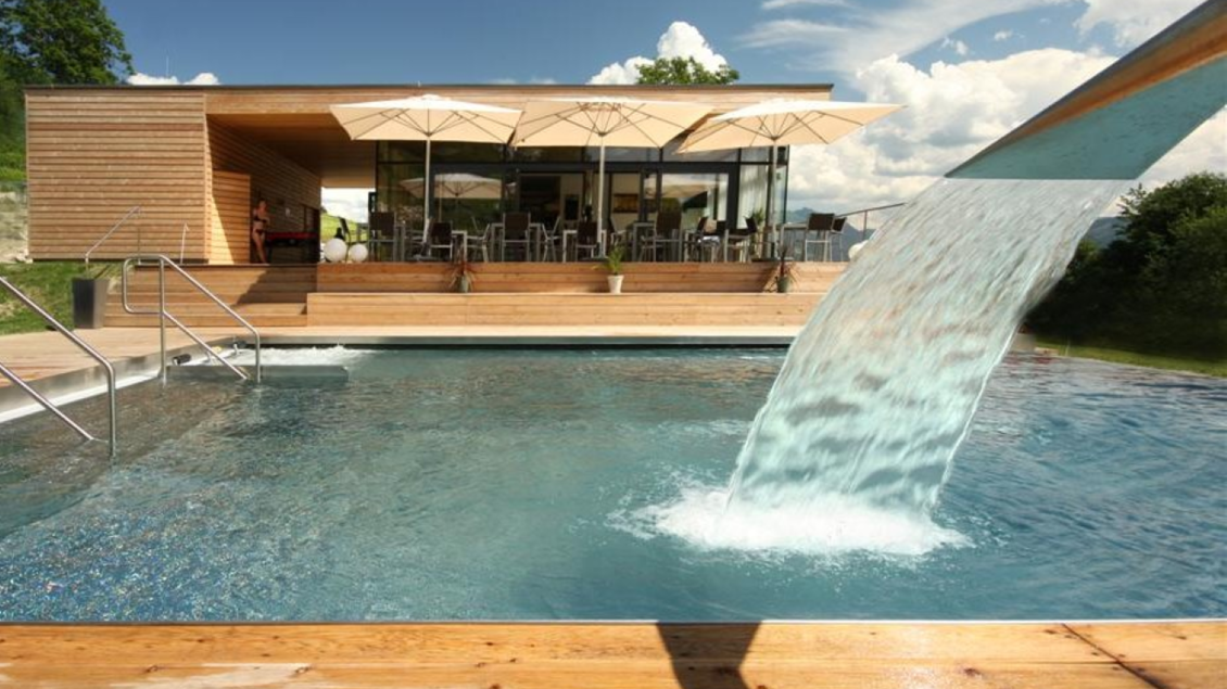 Das Bild zeigt ein modernes Naturbad mit klarem Pool, Holzdeck und einer Wasserfallrinne. Im Hintergrund lädt eine Terrasse mit Sonnenschirmen bei schönem Wetter zum Verweilen ein. | © Marktgemeinde Stainach-Pürgg - Kolb Roland
