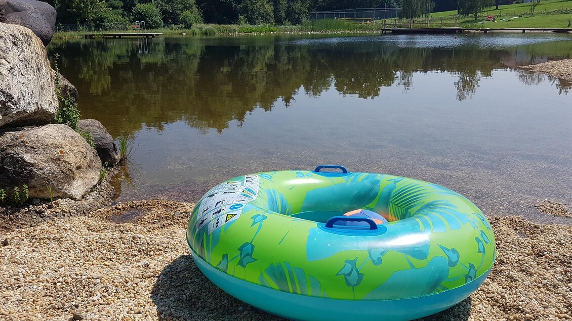 Ein bunter Schwimmreifen liegt am Ufer eines ruhigen Sees. Im Hintergrund sind Bäume und eine grüne Wiese zu sehen. | © Christine Schwetz