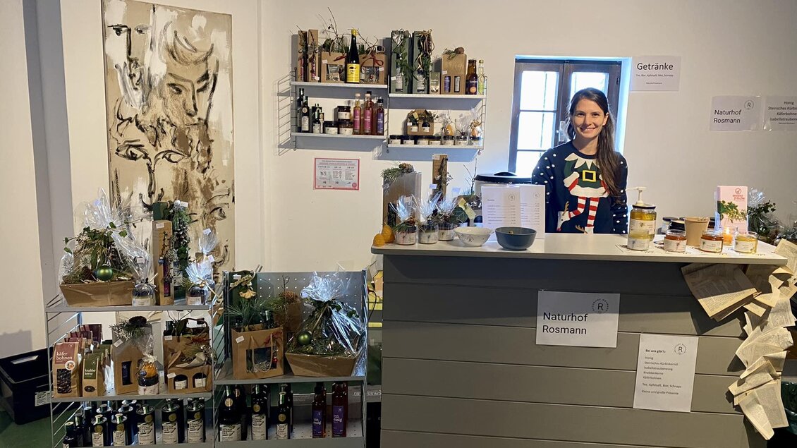 Ein Verkaufsstand mit Produkten auf Regalen und einer freundlichen Verkäuferin. Im Hintergrund ist ein Kunstwerk zu sehen. | © Imkerei Rosmann