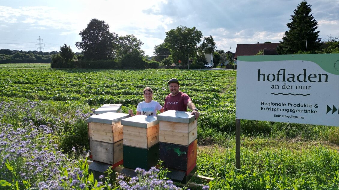Zwei Personen stehen neben Bienenstöcken auf einem Feld. Im Hintergrund ist ein Schild mit der Aufschrift "hofladen an der mur" zu sehen. | © Imkerei Rosmann