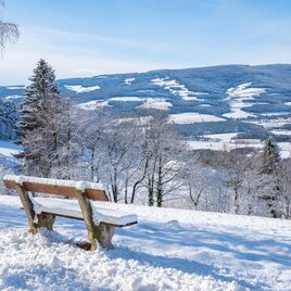 Eine verschneite Landschaft mit einem Blick auf Hügel und Täler. Eine Holzbank steht im Vordergrund, umgeben von Schnee und Bäumen. | © Helmut Schweighofer