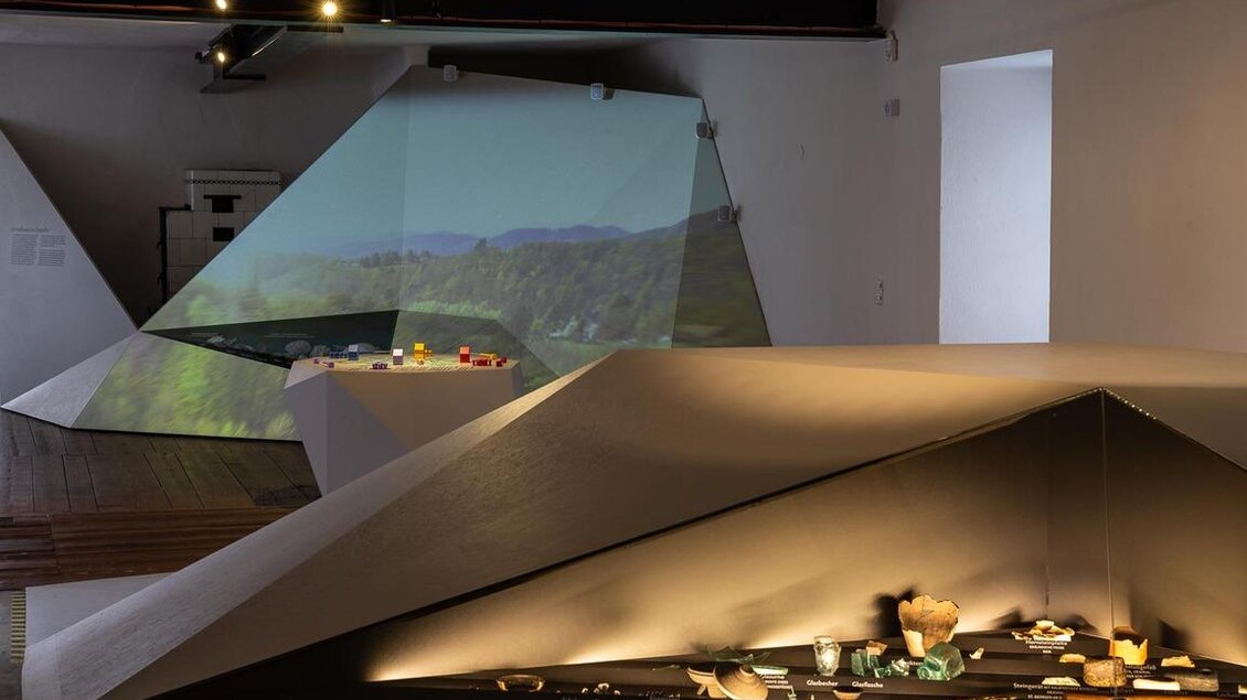 Ein moderner Ausstellungsraum mit geometrischen Strukturen. Auf einer Fläche sind Objekte und eine Projektion einer Landschaft zu sehen. | © Roland Marx - Naturparkzentrum Grottenhof