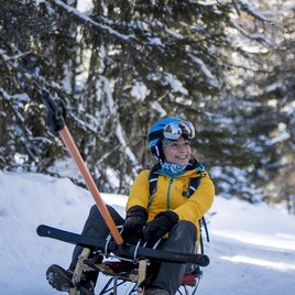 A person is sitting on a sled in the snow and wearing a helmet. In the background, there are snow-covered trees and a clear sky. | © Tom Lamm