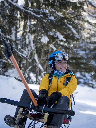 Eine Person sitzt auf einem Schlitten im Schnee und trägt einen Helm. Im Hintergrund sind verschneite Bäume und ein klarer Himmel zu sehen. | © Tom Lamm