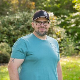 Ein Mann steht im Freien in einem grünen Park. Er trägt ein blaues T-Shirt und eine schwarze Kappe, mit Bäumen im Hintergrund. | © Helmut Schweighofer