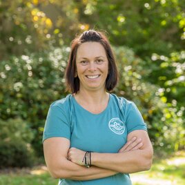 Eine lächelnde Frau mit kurzem, dunklem Haar steht in einem grünen Park. Sie trägt ein blaues T-Shirt und hat die Arme gekreuzt. | © Helmut Schweighofer