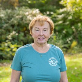 Eine ältere Frau steht auf einer Wiese mit Bäumen im Hintergrund. Sie trägt ein blaues T-Shirt und lächelt freundlich. | © Helmut Schweighofer